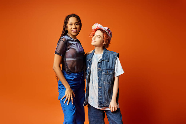 Дві молоді, одна небінарна, стоять об'єднані в стильний одяг на помаранчевому тлі в студійній обстановці. - Фото, зображення