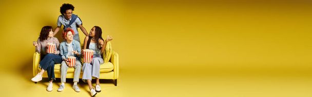Un grupo de jóvenes amigos multiculturales con un atuendo elegante sentados juntos en la parte superior de una silla amarilla en un entorno de estudio. - Foto, imagen