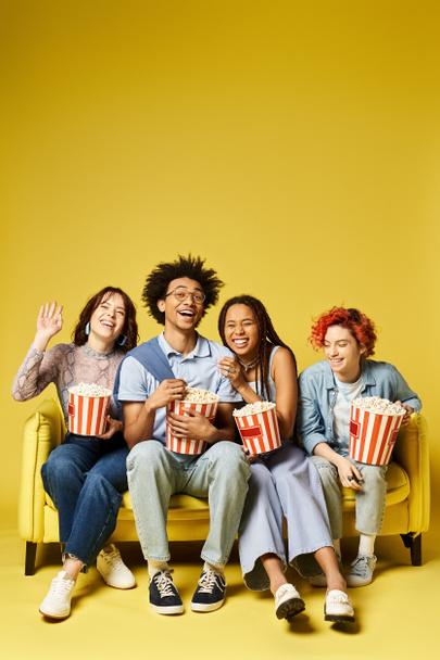Een groep jonge multiculturele vrienden, waaronder een niet-binaire persoon, zittend op een levendige gele bank in een stijlvolle studio-omgeving. - Foto, afbeelding