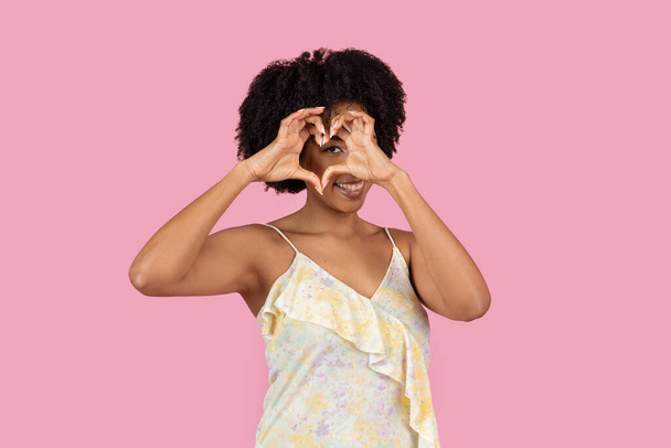 Una joven y confiada mujer afroamericana está de pie con un gesto en forma de corazón, sobre un fondo rosado vívido, con un vestido de verano amarillo - Foto, imagen