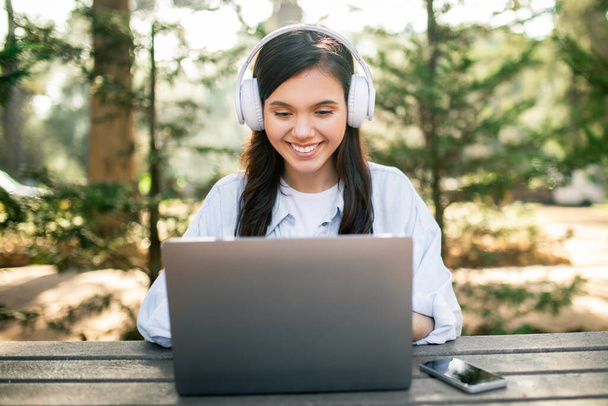 ヘッドフォンを持つ若いヨーロッパ人女性は,屋外の木製テーブルでラップトップを使用しながら笑顔,彼女の横のスマートフォン,外の緑豊かな森の背景に設定 - 写真・画像