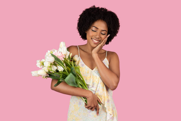 Eine junge Afroamerikanerin in einem floralen Kleid lächelt zärtlich, hält einen Strauß frischer Blumen vor rosa Hintergrund und verkörpert Glück und Dankbarkeit - Foto, Bild