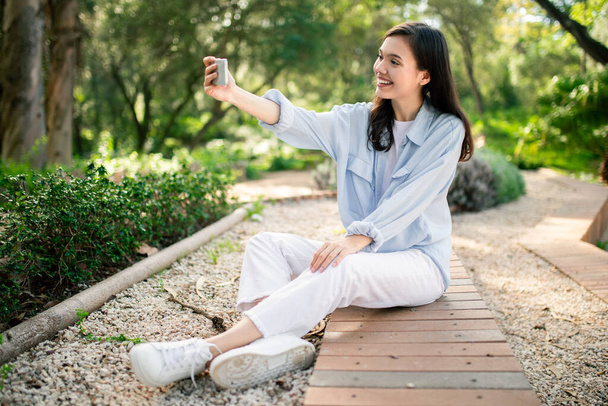 Hevesli bir kadının parkta akıllı telefonuyla kendi fotoğrafını çekmesi sosyal paylaşım zevkini resmediyor. - Fotoğraf, Görsel