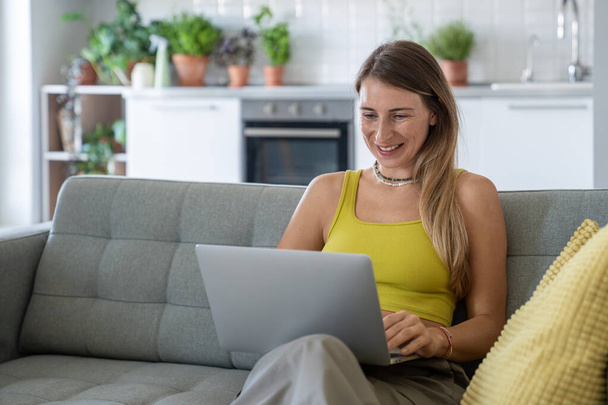 Szczęśliwa kobieta siedząca z laptopem na kanapie otrzymuje ciekawe dobre wieści. Zadowolona wesoła kobieta z przenośnym komputerem na kolanach na czacie w mediach społecznościowych, zakupy online, baw się dobrze w internecie. - Zdjęcie, obraz