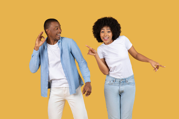Glimlachende Afro-Amerikaanse man en vrouw casual gekleed, dansen en wijzen, genieten op een gele achtergrond - Foto, afbeelding