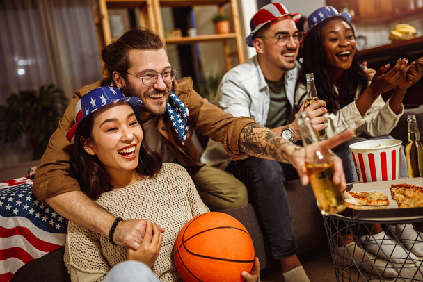Eine multiethnische Gruppe von Freunden beobachtet Basketballspiel mit amerikanischen Flaggen, trinkt Bier und jubelt. - Foto, Bild