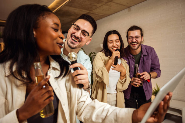Grupo multirracial de amigos teniendo una noche de karaoke. Cantando en el micrófono, mirando letras y bebiendo cerveza. - Foto, Imagen
