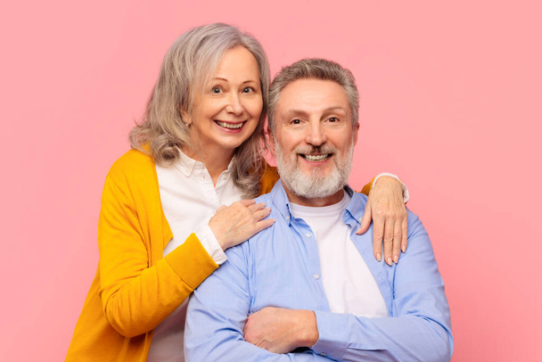 Eine Ehe in Reife. Fröhliches älteres Paar, das Glück und Romantik ausstrahlt, sich vor rosa Studiokulisse umarmt und in die Kamera lächelt. Porträt verliebter Senioren, die gemeinsam posieren - Foto, Bild