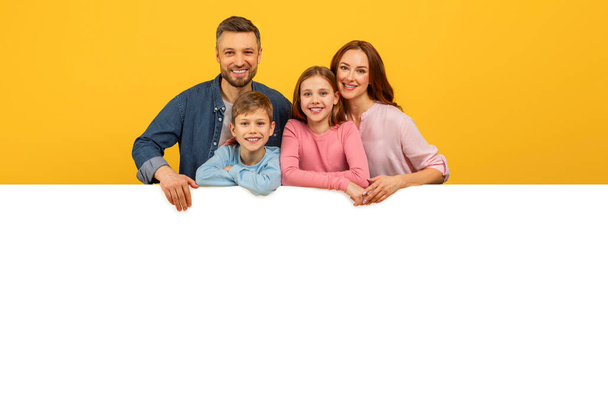 Mosolygó család kukucskál ki egy üres fehér tábla felett, tökéletes reklámok vagy másolás tér mockup sárga háttér - Fotó, kép