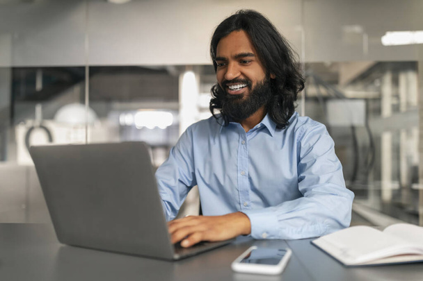 Uvolněný profesionální muž s lehkým úsměvem pracuje na svém notebooku v pohodlném kancelářském prostředí - Fotografie, Obrázek