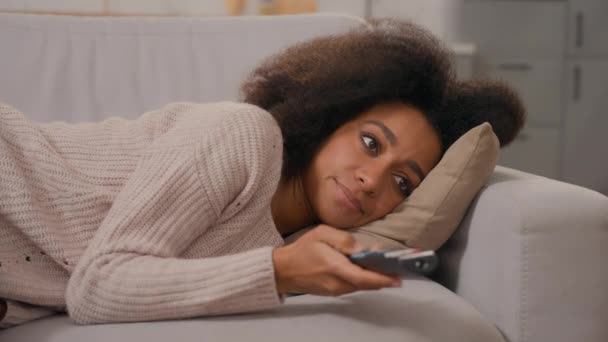 Znudzony niezadowolony zdenerwowany Afroamerykanka samotna młoda dziewczyna leniwy właściciel domu kobieta leżąca na kanapie kanapy wyszukiwania programu telewizyjnego z pilotem zdalnego sterowania oglądania telewizji nudne filmy w domu - Materiał filmowy, wideo
