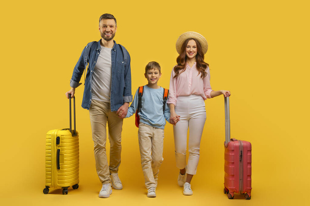 Familia sonriente de tres, padres e hijo, que sostiene maletas contra un fondo amarillo, listo para viajar de vacaciones - Foto, Imagen
