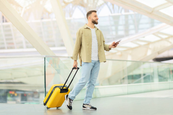 Un homme à la mode se promène avec une valise jaune vif tout en utilisant son téléphone à l'aéroport - Photo, image