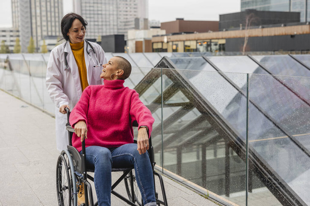 Een moment van echte verbondenheid als een lachende arts communiceert met haar patiënt in een rolstoel tegen een stedelijke achtergrond. - Foto, afbeelding