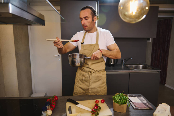ハンサムな白人男性は,現代の家庭用キッチンでストーブに立ってトマトソースを味わいます. 伝統的なイタリアのレシピに従って新鮮な有機トマトとソースを調理する男性シェフ. - 写真・画像