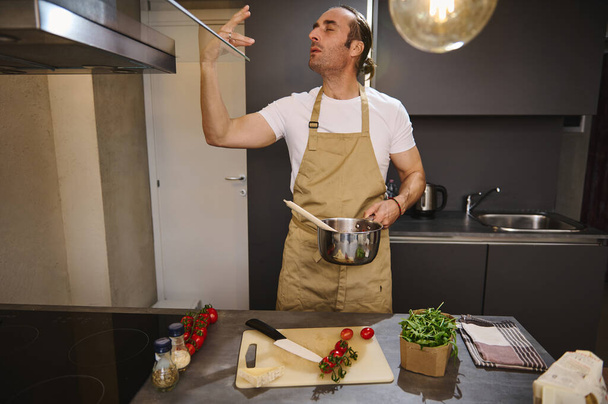 Handsome male chef holding saucepan with tomato sauce, kissing fingers, showing tasty hands sign, saying bellissimo cooking gourmet food in the home kitchen. People. Culinary. Alimentation. Lifestyle. - Photo, Image