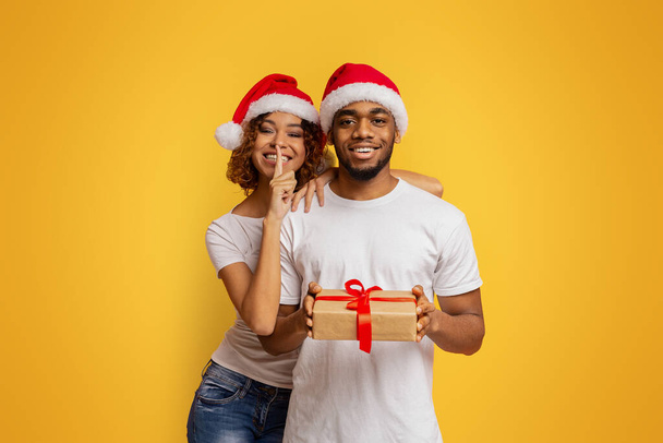 Christmas surprise. Millennial african-american couple in Santa hats holding Xmas gift, girl showing silence sign, orange background - Photo, Image