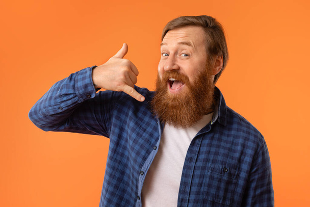 Веселый рыжеволосый бородатый мужчина жестикулирует Call Me рукой возле уха, притворяясь, что он отвечает на телефонный звонок, на фоне оранжевой студии, портретный снимок. Концепция телефонного номера и связи - Фото, изображение