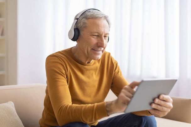 Starszy mężczyzna skupia się na tablecie ze słuchawkami, wskazując, że może cieszyć się multimediami lub uczyć się czegoś nowego. - Zdjęcie, obraz