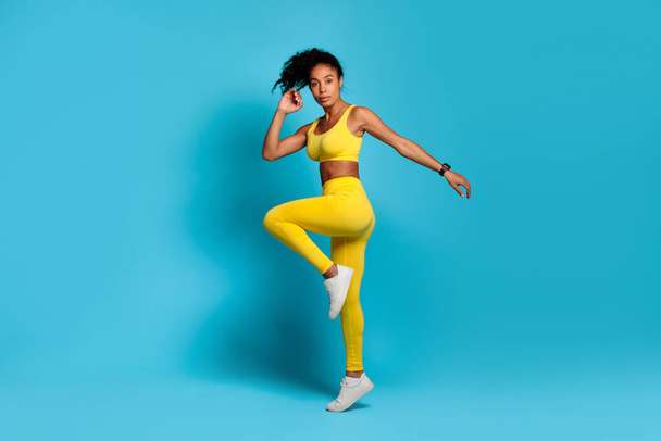 Fit dame afro-américaine dans l'athlétisme jaune frappant une pose d'équilibre ludique sur un fond bleu solide, montrant la flexibilité - Photo, image