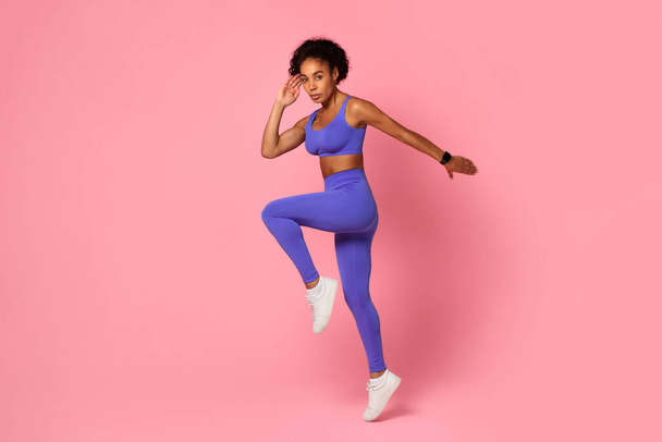 Una animada mujer afroamericana en ropa deportiva azul haciendo ejercicio con una pierna levantada, exudando confianza sobre un telón de fondo rosa - Foto, Imagen