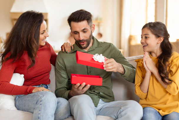 Nadšený otec rozbaluje červený dárek se svou rodinou, zachycuje okamžik radosti a překvapení na gauči - Fotografie, Obrázek