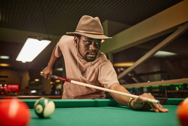 Retrato dramático del hombre afroamericano adulto jugando al billar y golpeando la pelota con palo en el espacio de copia del club de billar - Foto, Imagen