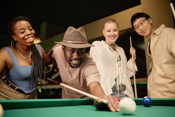 Multiethnische Gruppe von Freunden spielen Pool zusammen mit Fokus auf lächelnde schwarze Mann schlagen Ball mit Cue-Stick Kopierraum - Foto, Bild