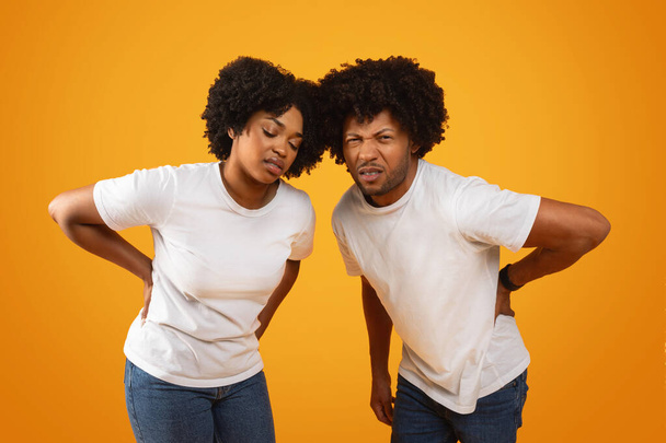 Unhappy millennial african american spouses suffer from lower back pain. Sick tired black man and woman touching their back, yellow background. Bad posture, osteoporosis, muscle strain - Photo, Image