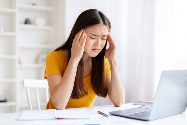 Jovem mulher asiática sentindo estresse e dor de cabeça enquanto trabalhava no laptop em casa escritório, esfregando seus templos - Foto, Imagem