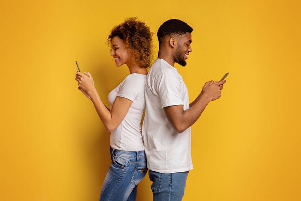 Ένα νεαρό ζευγάρι Αφροαμερικανών στέκεται πλάτη με πλάτη, απορροφημένο στα smartphones τους, συμβολίζοντας σύγχρονα θέματα επικοινωνίας σε ένα ζωντανό κίτρινο φόντο - Φωτογραφία, εικόνα