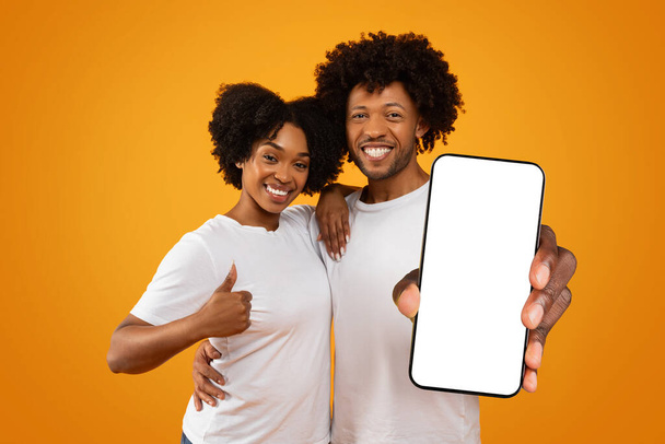 Liebendes schwarzes Millennial-Paar zeigt großes Handy mit weißem Leerbildschirm-Kopierplatz für mobile App-Werbung, Online-Angebot, isoliert auf gelbem Studiohintergrund - Foto, Bild