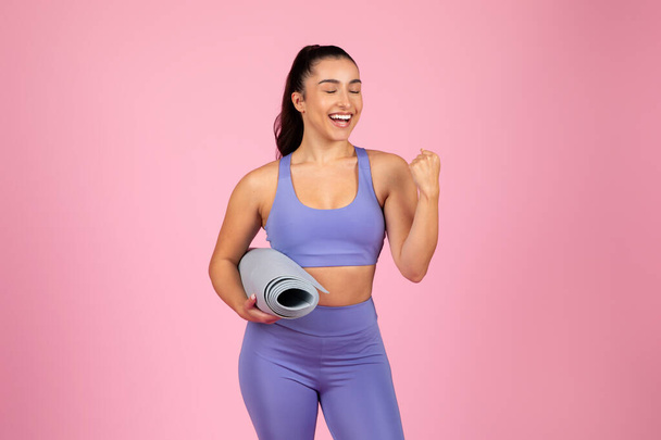Eine energische Frau in Trainingsausrüstung hält eine zusammengerollte Yogamatte in der Hand, lächelt auf rosa Hintergrund und strahlt Glück aus. - Foto, Bild