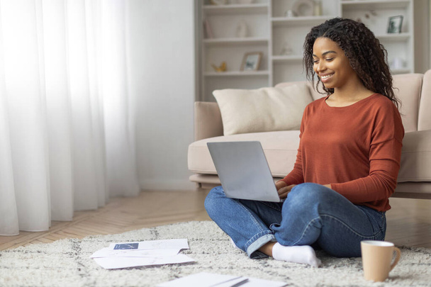 Schwarze Frau sitzt auf dem Boden und konzentriert sich auf einen Laptop-Bildschirm. afrikanisch-amerikanische Frau, die von zu Hause aus online arbeitet - Foto, Bild