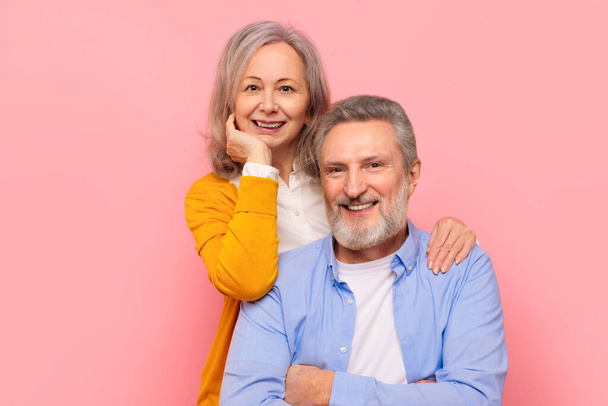 Ältere Liebe. Lächelndes Senioren-Paar, das sich umarmt und für ein Familienporträt posiert, die Wärme ihrer anhaltenden romantischen Beziehung genießt, zusammen vor rosa Studiokulisse steht und in die Kamera blickt - Foto, Bild