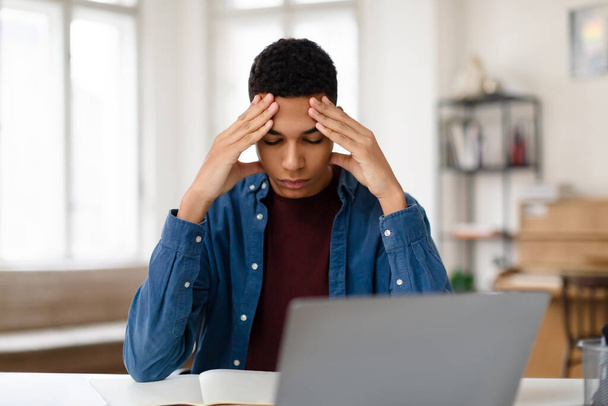 Enfocado chico adolescente negro que experimenta dolor de cabeza mientras se concentra en sus estudios delante de la computadora portátil, concepto de estrés estudiantil y presión académica - Foto, imagen