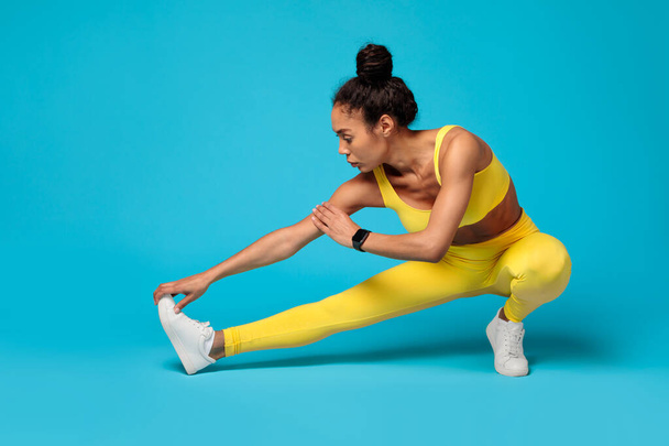 Συγκεντρωμένη Αφροαμερικανίδα αθλήτρια σε ζωηρά κίτρινα αθλητικά ρούχα τεντώνεται κατά τη διάρκεια της προπόνησής της σε ένα μπλε φόντο - Φωτογραφία, εικόνα