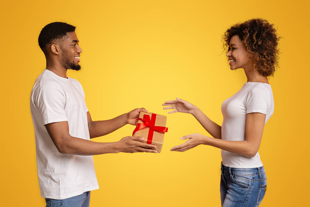 Afro-Amerikaanse jongeman presenteert een rood lint verpakt geschenkdoos aan een opgetogen jonge vrouw tegen een gele achtergrond - Foto, afbeelding