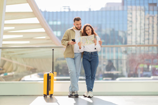 Любляча пара тримає карту паперу і використовує телефон, стоячи в аеропорту з жовтою валізою - Фото, зображення
