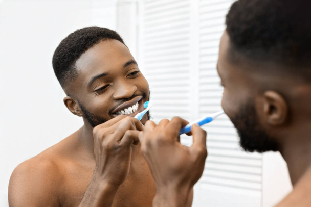 Afrykański Amerykanin bez koszulki myje zęby w lustrze, reprezentując dobre nawyki higieniczne i pielęgnacyjne. - Zdjęcie, obraz