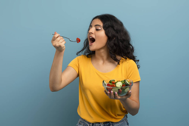A fiatal nő élvezi a friss salátát, vidám kifejezéssel, kék háttérrel. - Fotó, kép