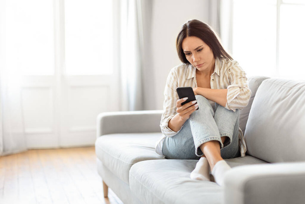 Frau sieht nachdenklich und nachdenklich aus, als sie zu Hause in einem lichtdurchfluteten Raum auf einer Couch sitzt und ein Smartphone in der Hand hält - Foto, Bild