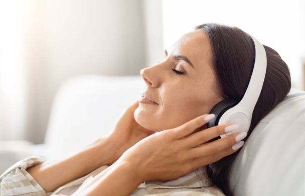 Una mujer exhausta descansando en un sofá, con los auriculares puestos, que representa la relajación con un toque de tecnología - Foto, Imagen