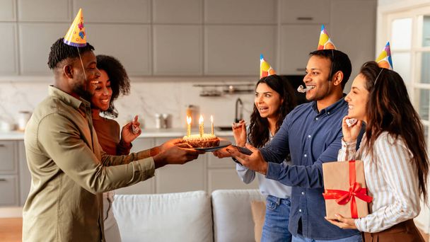 Des amis multiraciaux tenant un gâteau d'anniversaire avec des bougies allumées le présentent à une femme souriante lors d'une célébration joyeuse - Photo, image