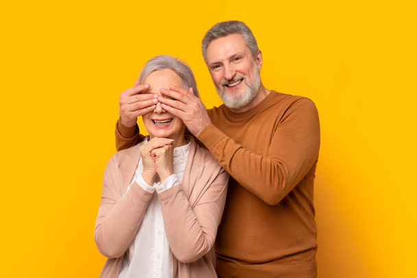 Allegro uomo anziano europeo chiude gli occhi a sua moglie sorridente alla macchina fotografica, preparando anniversario o sorpresa di compleanno, in piedi su sfondo giallo studio. Sorprese romantiche, Celebrazione - Foto, immagini