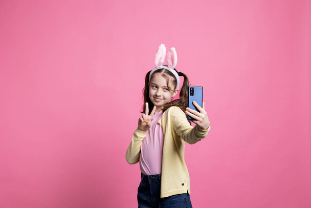 Radosne szczęśliwe dziecko robi zdjęcia i pokazuje znak pokoju przez telefon, czując się wesoły i pewny siebie o zabawnych zdjęć do obchodów Wielkanocy. Mała uczennica z warkoczami robi zdjęcia. - Zdjęcie, obraz