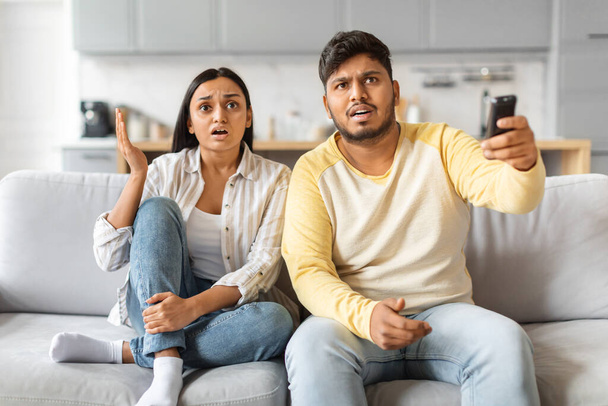 Indian couple seated on a couch displays shock and tension while watching television together, young couple emotionally reacting to content - Photo, Image