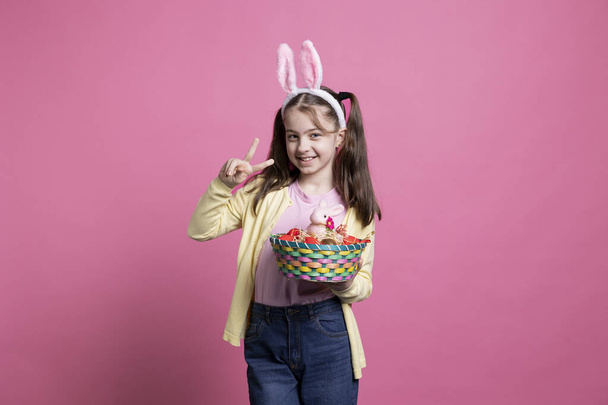 Маленька дівчинка демонструє Великодній дух, тримаючи чарівні іграшки в студії, демонструючи знак миру перед камерою. Маленька дитина з вухами кролика і косичками позує з кошиком для яєць і кроликом. - Фото, зображення
