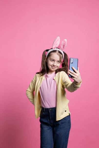 Positive cute schoolgirl taking pictures with her mobile phone in studio, smiling at camera and posing with confidence against pink background. Cheerful young kid takes photos for easter festivity. - Photo, Image