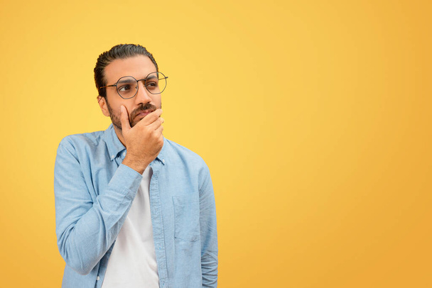Pensive indian man in a denim shirt poses thoughtfully with hand on chin against a uniform yellow background - Photo, Image